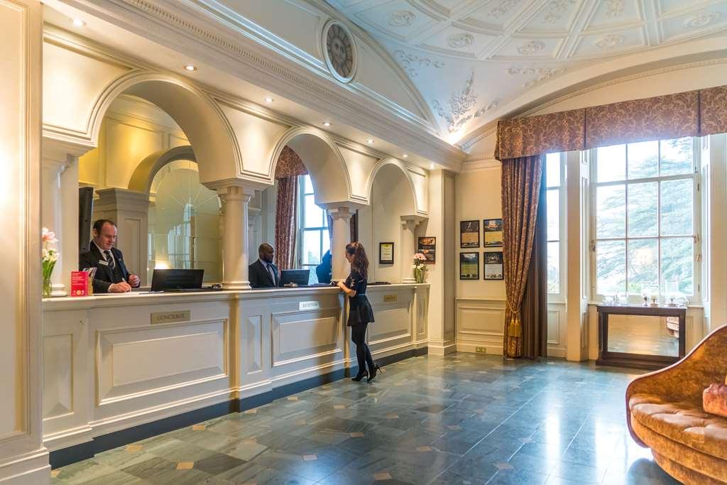 Macdonald Bath Spa Hotel Wnętrze zdjęcie The photo shows the interior of a hotel reception area. There is a stylish front desk with several staff members attending to guests. A woman is interacting with one of the receptionists. The design features elegant architectural details, including a