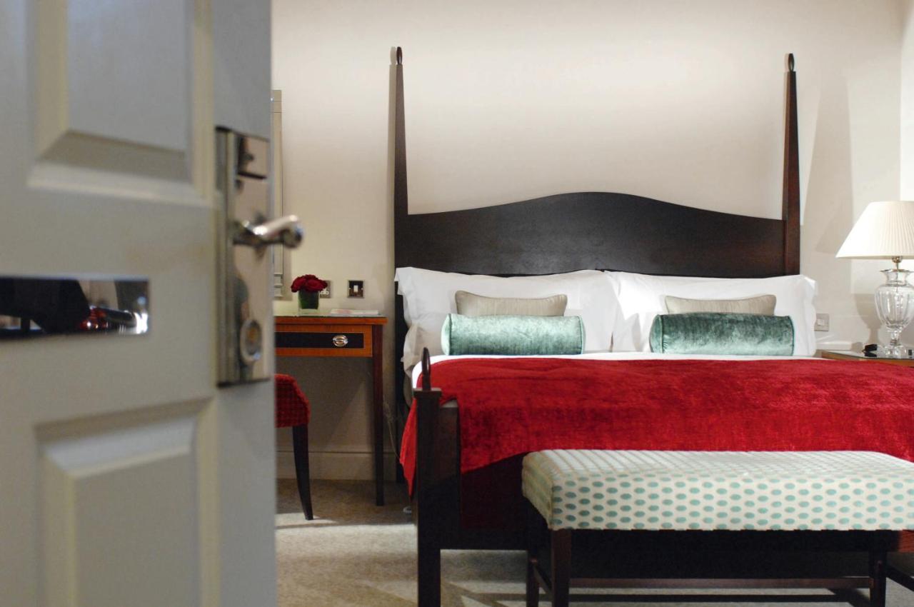 Macdonald Bath Spa Hotel Pokój zdjęcie The photo shows a well-decorated hotel room. In the foreground, there is a partially opened door leading into the room. The focal point of the room is a large bed with a dark wooden headboard and stylish red and white bedding, accented with green pil