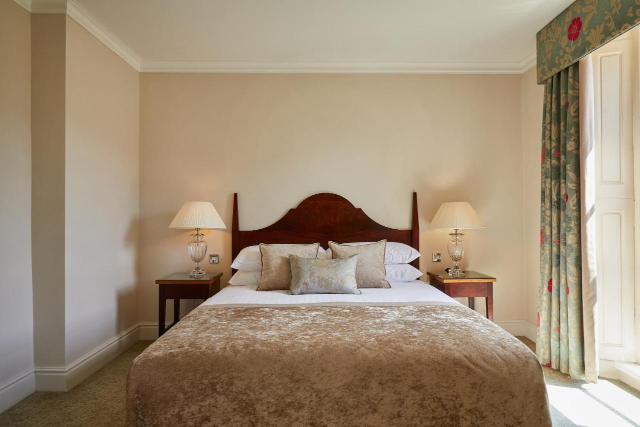 Macdonald Bath Spa Hotel Zewnętrze zdjęcie The photo shows a cozy hotel bedroom. It features a large bed with a plush, light-colored bedspread and several pillows. On either side of the bed, there are wooden nightstands with lamps providing soft lighting. The walls are painted a neutral color