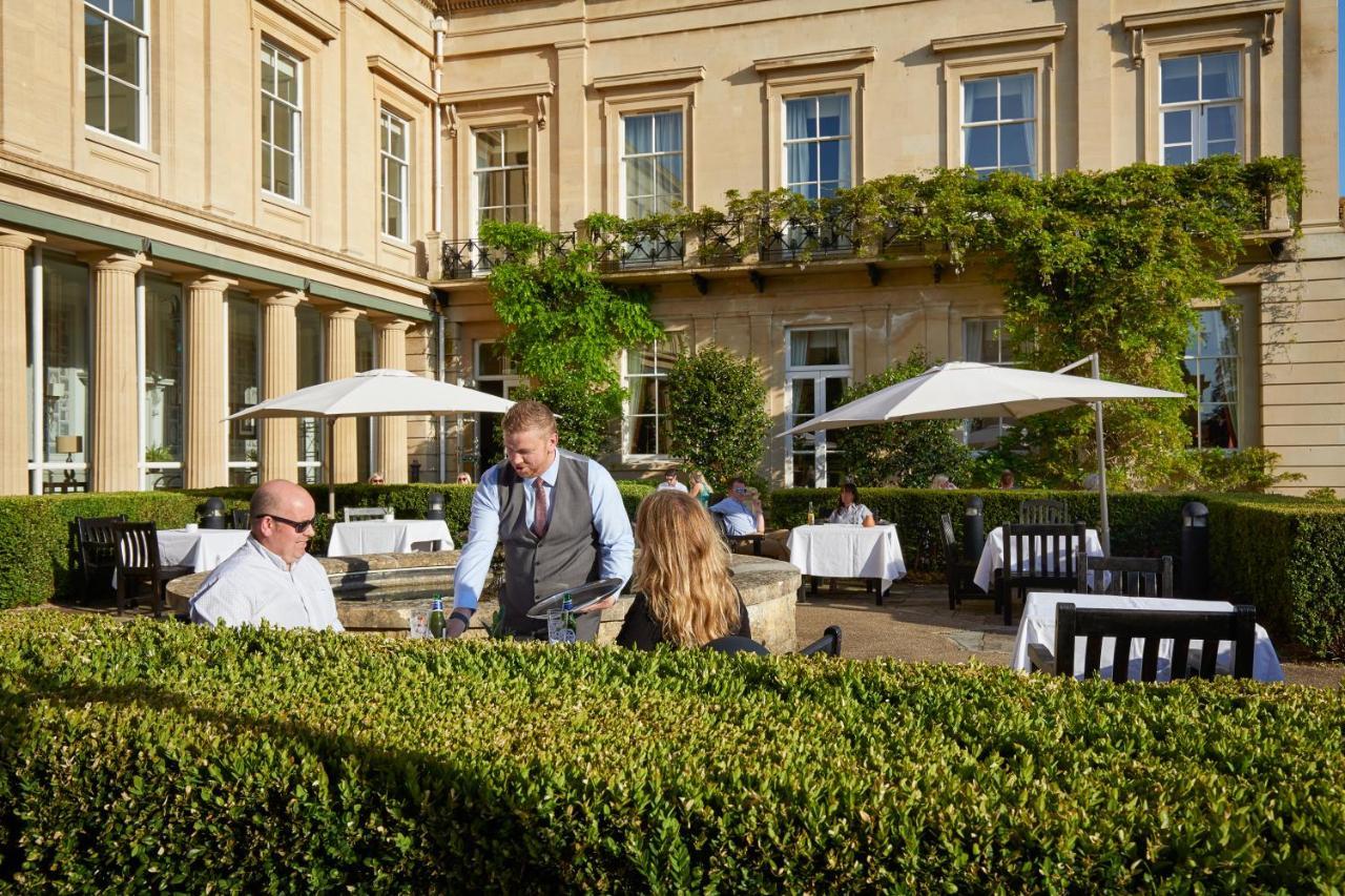 Macdonald Bath Spa Hotel Zewnętrze zdjęcie The photo depicts an outdoor dining area of a restaurant. In the foreground, there are neatly trimmed hedges surrounding the dining space. Two patrons are seated at a table; one is a man wearing sunglasses and the other is a woman facing him. A waite