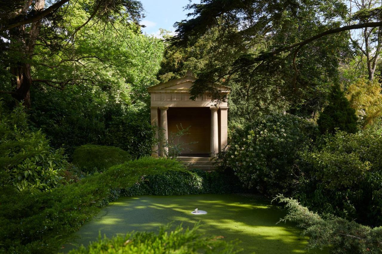 Macdonald Bath Spa Hotel Zewnętrze zdjęcie The photo shows a serene garden scene featuring lush greenery and a tranquil pond. In the background, there is a classical-looking structure with columns, resembling a small temple or pavilion. The pond appears to have vegetation covering its surface