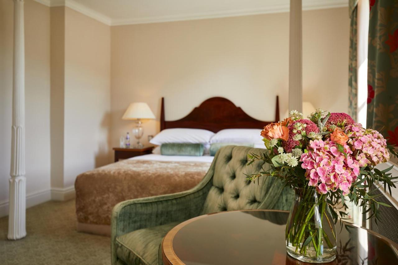 Macdonald Bath Spa Hotel Zewnętrze zdjęcie The photo shows a cozy bedroom featuring a large bed with a wooden headboard, topped with a fluffy blanket. To one side, there is a small bedside lamp providing soft lighting. In the foreground, there is an elegant green chair with a tufted design an