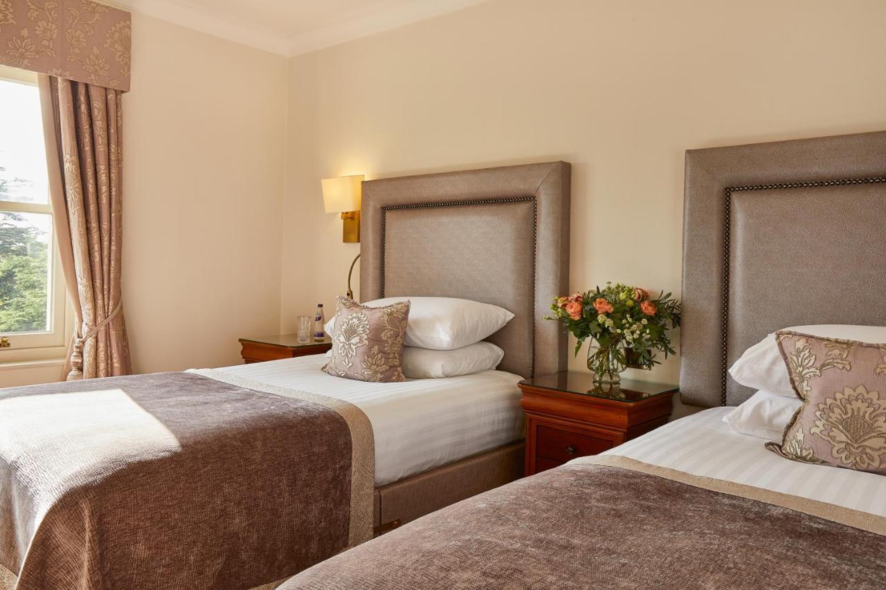 Macdonald Bath Spa Hotel Zewnętrze zdjęcie The photo shows a hotel room featuring two beds with elegant headboards. Each bed is neatly made with white linens and a soft, decorative throw blanket. There are matching pillows on the beds, and a small, stylish bouquet of flowers is placed on a wo