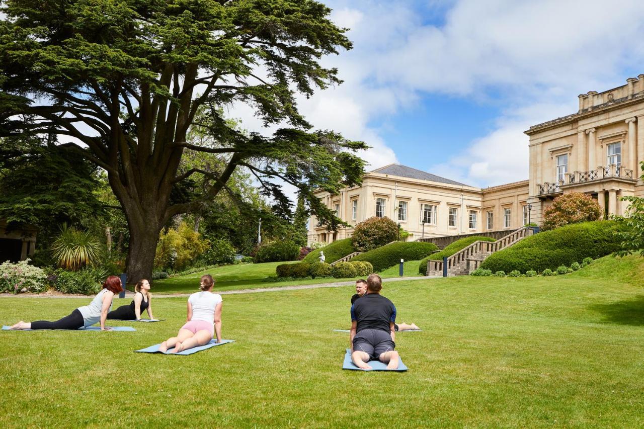 Macdonald Bath Spa Hotel Zewnętrze zdjęcie The photo shows a group of people engaging in a yoga session outdoors. They are positioned on yoga mats on a lush green lawn in front of a large, elegant building with columns. Behind them, there are trees and well-maintained gardens, contributing to