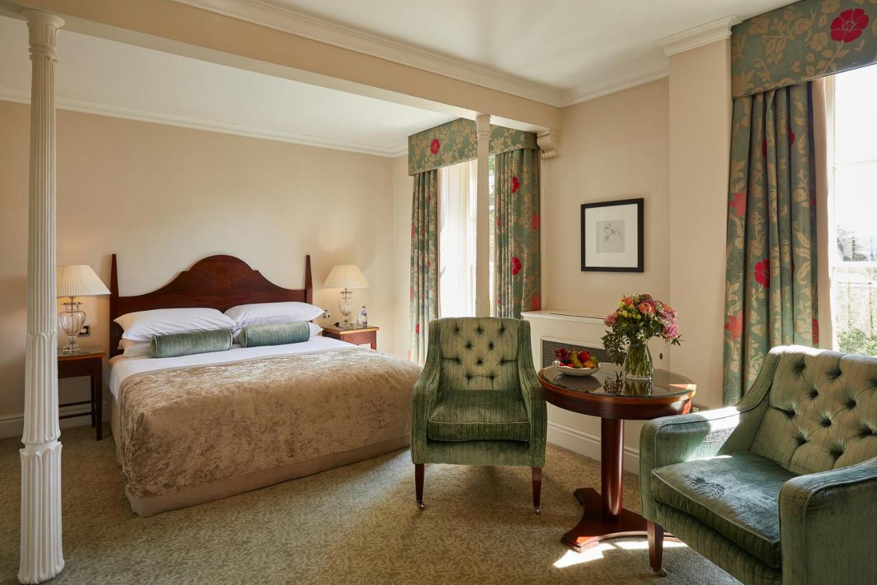 Macdonald Bath Spa Hotel Zewnętrze zdjęcie The photo shows a cozy hotel room featuring a large bed with a beige bedspread. There are two green armchairs positioned next to a small round table. The decor includes floral curtains and a neutral color palette on the walls. Soft lighting is provid