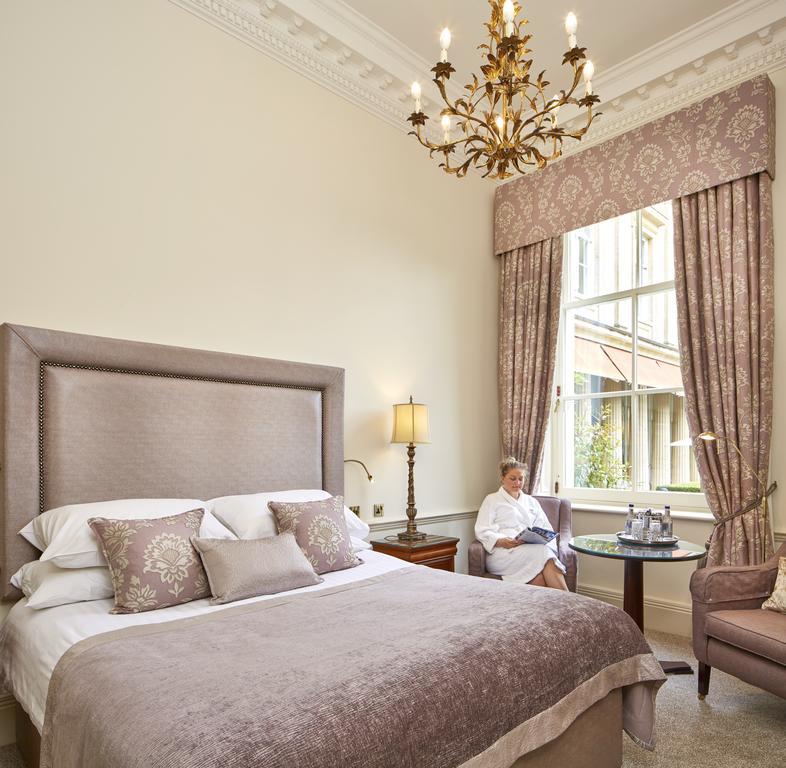 Macdonald Bath Spa Hotel Zewnętrze zdjęcie The photo shows a well-appointed hotel room that features a large, elegant bed with soft bedding and decorative pillows. The walls are painted a light color, and there is an ornate chandelier hanging from the ceiling. A beautiful window is adorned wi