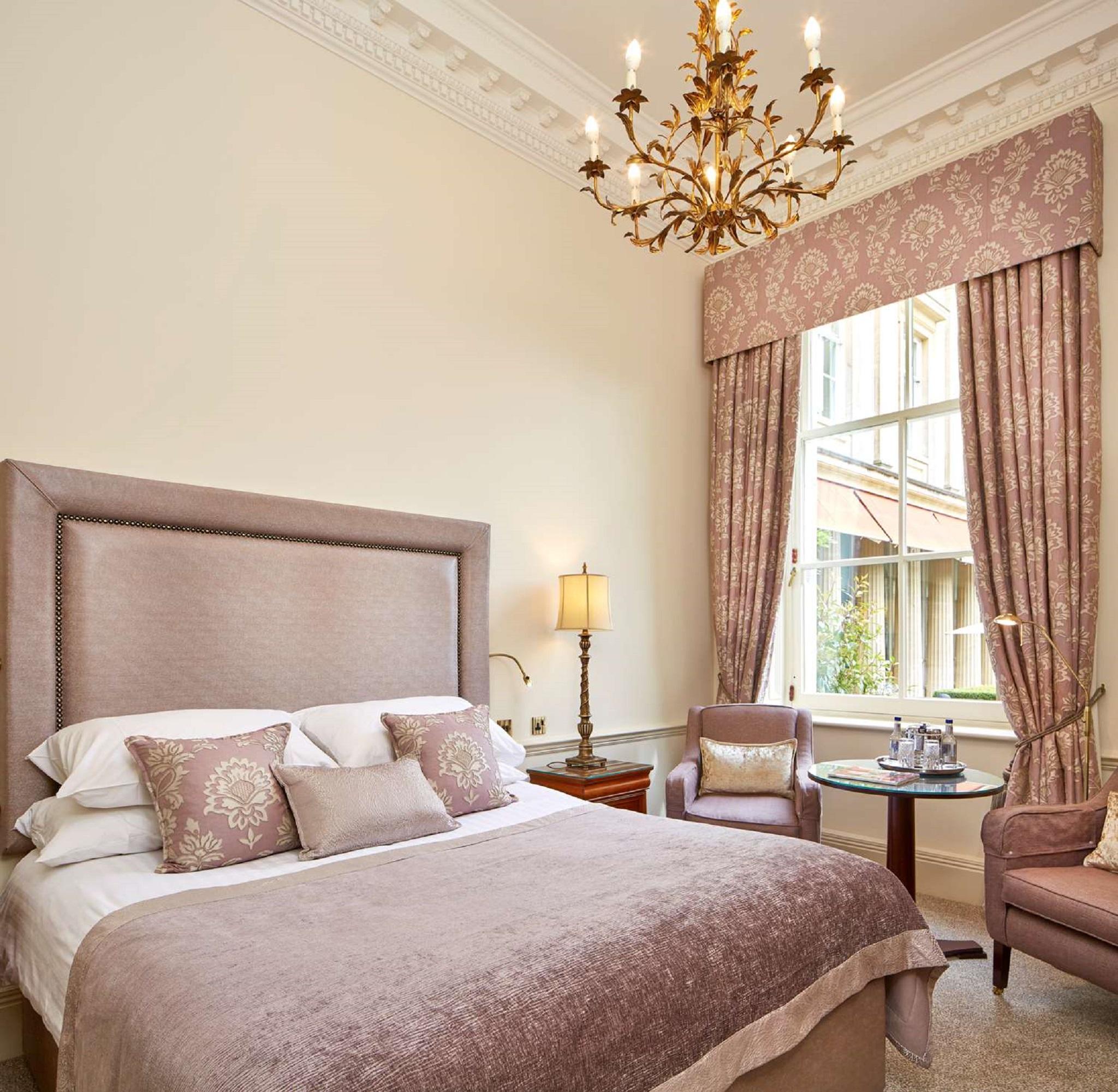 Macdonald Bath Spa Hotel Zewnętrze zdjęcie The photo shows a beautifully decorated hotel room. It features a large bed with a plush headboard, adorned with several decorative pillows. The walls are painted in a soft color, and there is a chandelier hanging from the ceiling. Natural light come