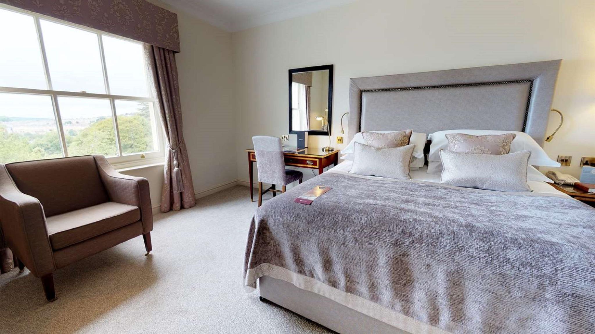 Macdonald Bath Spa Hotel Zewnętrze zdjęcie The photo shows a cozy and well-decorated bedroom. It features a large bed with a plush, grey bedspread and several decorative pillows. Beside the bed, there is a wooden bedside table with a lamp. A small desk and chair are positioned near a window, 