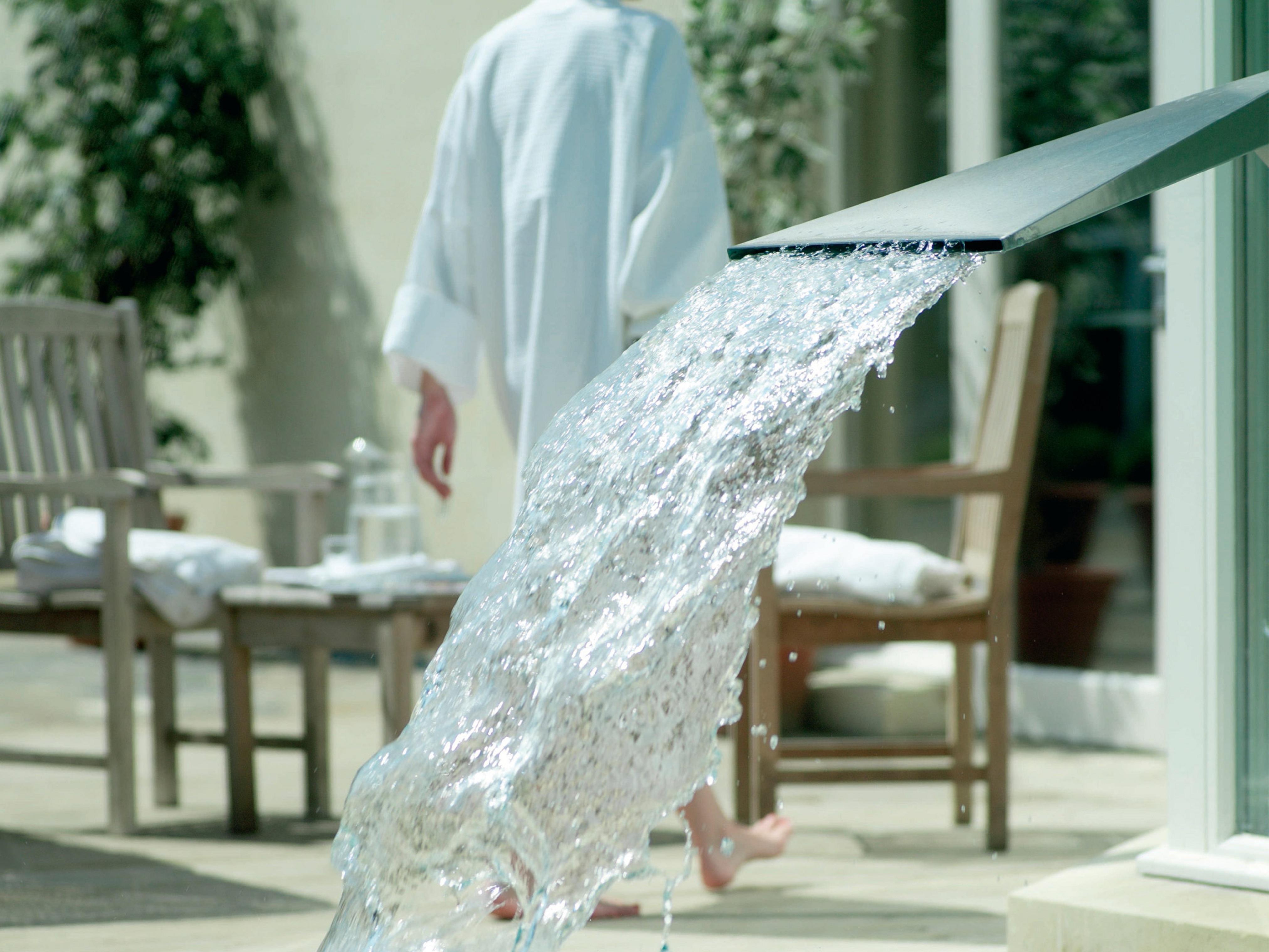 Macdonald Bath Spa Hotel Zewnętrze zdjęcie The photo shows a flowing water feature, with water elegantly cascading from a sleek spout. In the background, there is a person wearing a white bathrobe, walking away. Surrounding the area, there are wooden chairs and what appears to be a serene out