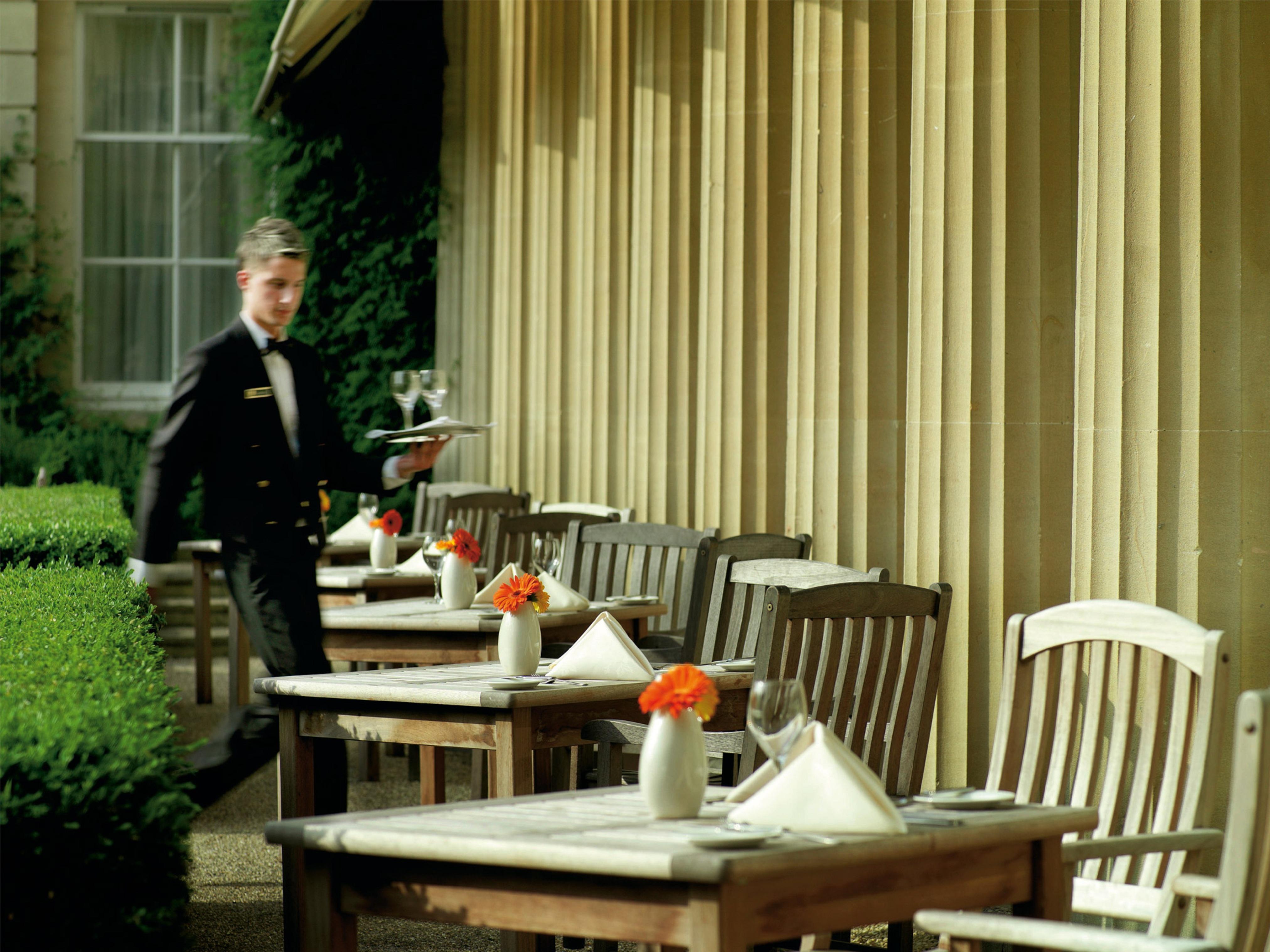 Macdonald Bath Spa Hotel Zewnętrze zdjęcie The photo shows an outdoor dining area, featuring several wooden tables and chairs arranged neatly. Each table is set with a white tablecloth and small vases containing flowers, adding a vibrant touch. A waiter in a formal black suit is seen moving t