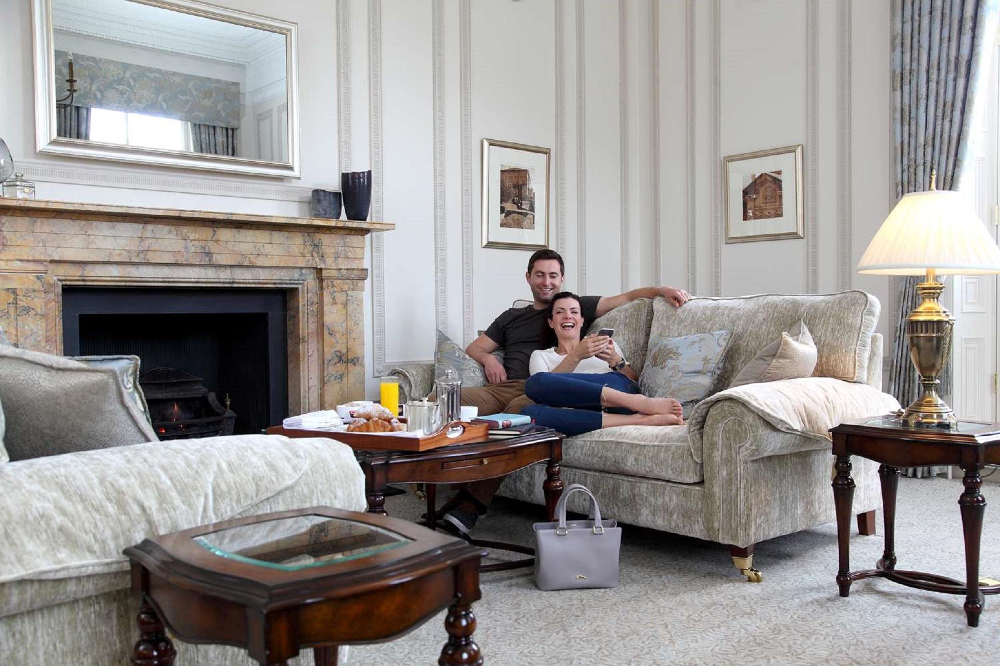Macdonald Bath Spa Hotel Zewnętrze zdjęcie The photo shows a cozy living room setting where a couple is sitting together on a light-colored sofa. They appear relaxed, with the man sitting slightly behind the woman, who is smiling and looking at her phone. The room features elegant decor, incl
