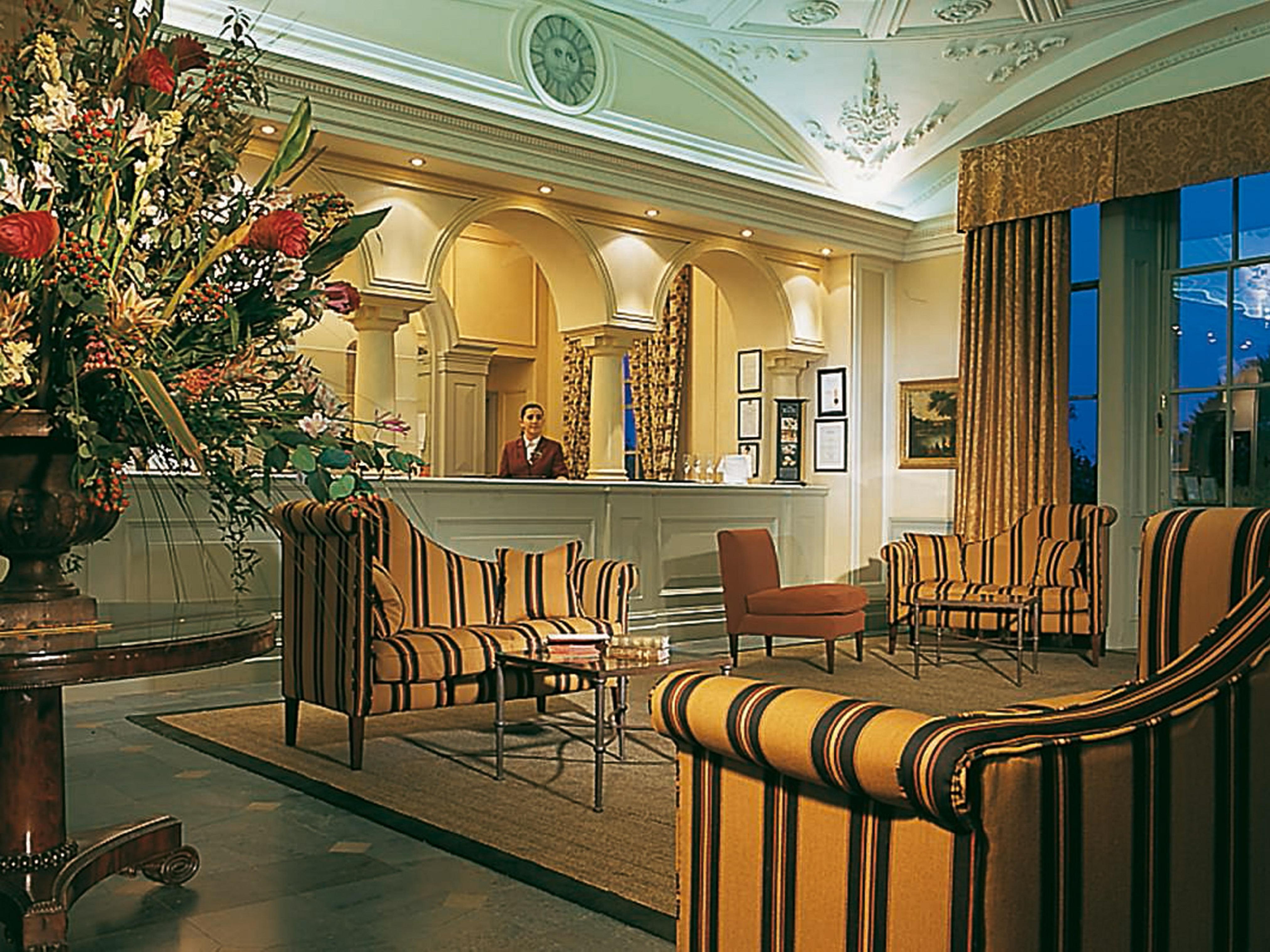 Macdonald Bath Spa Hotel Zewnętrze zdjęcie The photo shows an elegant hotel lobby. The area features a reception desk with a staff member behind it. Surrounding the reception are comfortable seating arrangements, including striped armchairs and a brown chair. A large vase with an elaborate fl