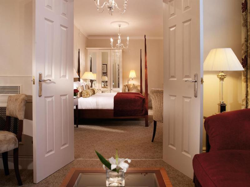 Macdonald Bath Spa Hotel Pokój zdjęcie The photo shows a hotel room that can be viewed through open double doors. Inside, there is a neatly made bed with decorative pillows, a plush area rug, and soft lighting from lamps. The room features elegant furnishings, including a chair, a coffee 