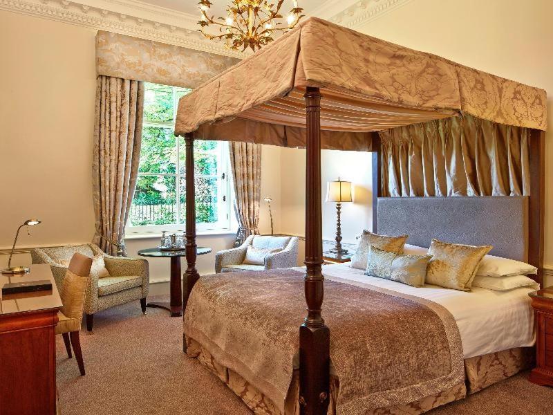 Macdonald Bath Spa Hotel Pokój zdjęcie The photo shows an elegantly designed bedroom featuring a four-poster bed with a decorative canopy. The bed is dressed with luxurious bedding and pillows, creating a cozy and inviting atmosphere. Flanking the bed are two comfortable armchairs and a s