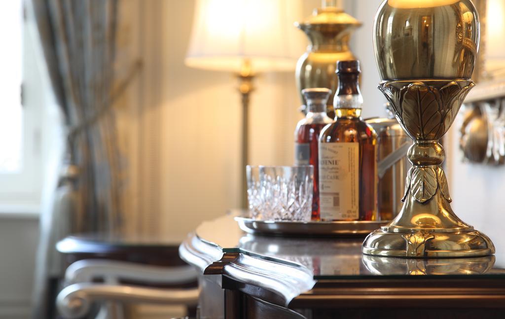 Macdonald Bath Spa Hotel Zewnętrze zdjęcie The photo features an elegant interior scene that includes a polished wooden bar or table. On the surface, there are several bottles of liquor, likely whiskey, each with a distinctive label. A glass, possibly for drinking, is positioned near the bott